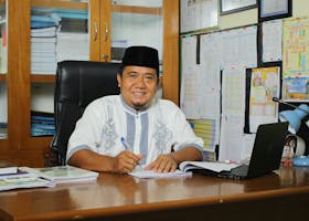 An Indonesian teacher in traditional attire, smiling at his desk with documents in an office setting.